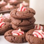 Peppermint Kiss Cookies are chocolate cookies topped with red and white striped peppermint candies, stacked next to a glass of milk.