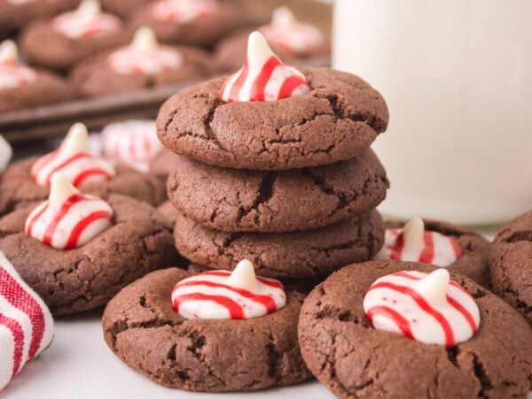 Peppermint Kiss Cookies are chocolate cookies topped with red and white striped peppermint candies, stacked next to a glass of milk.