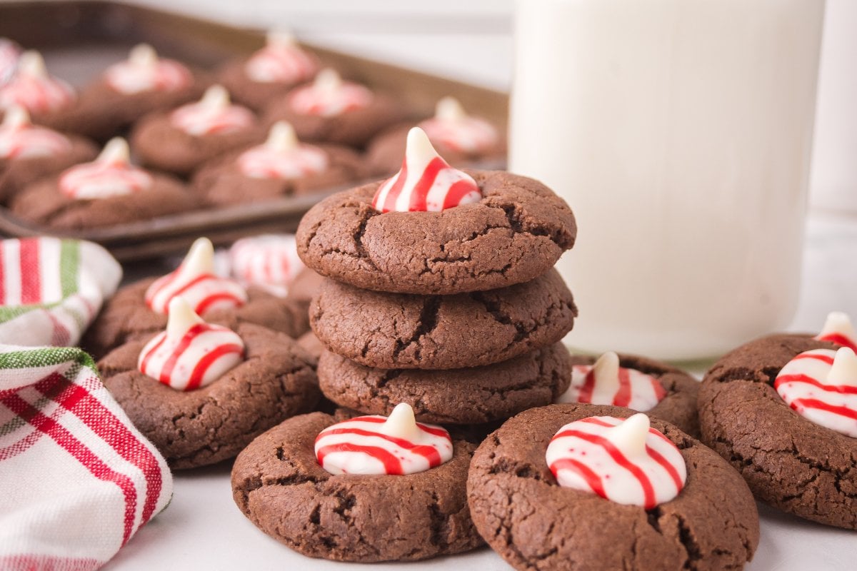 Peppermint Kiss Cookies: Chocolate cookies with red and white peppermint candies, stacked by a glass of milk and a striped cloth.