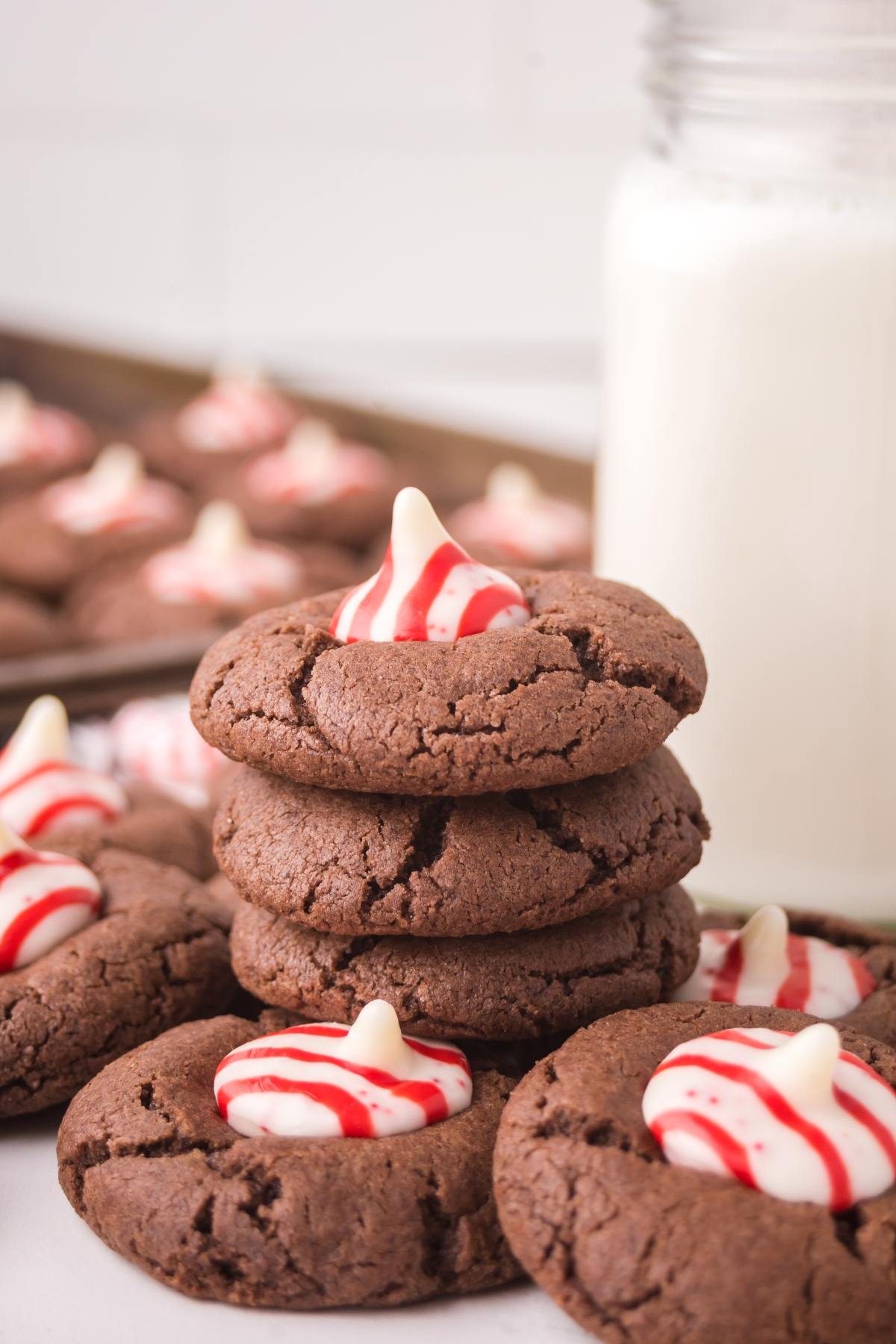 A stack of chocolate Peppermint kiss cookies topped with red and white striped candies, with a glass of milk in the background.