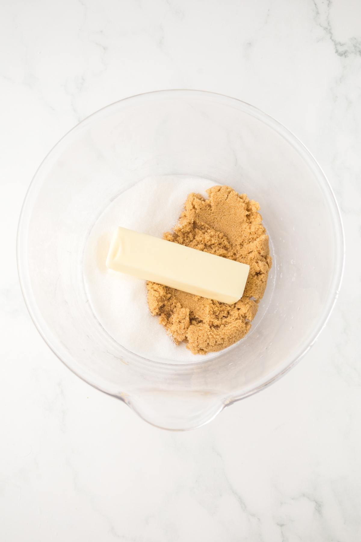A glass bowl with butter, granulated sugar, and brown sugar on a white marble surface.