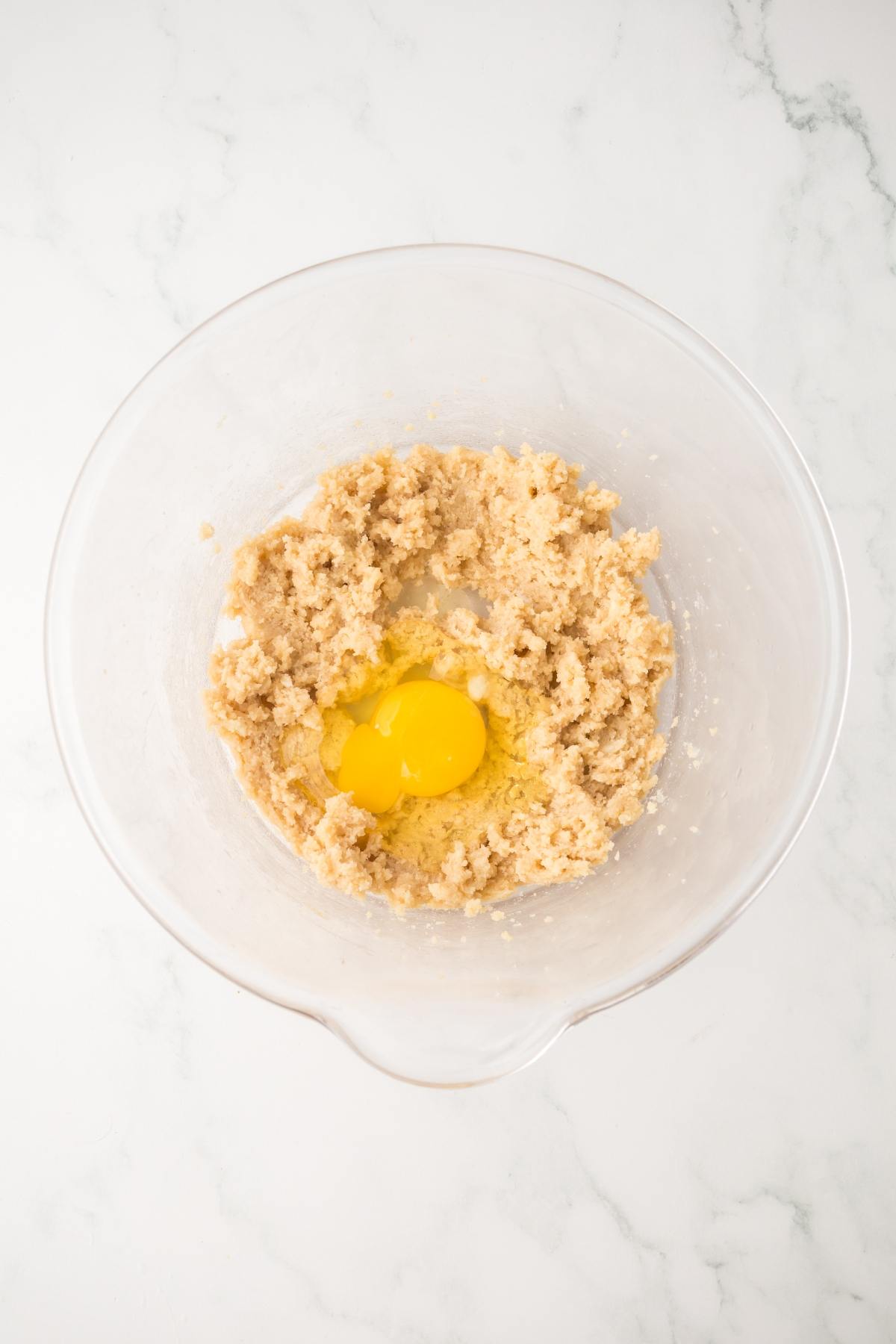 A mixing bowl with creamed butter and sugar, and an egg cracked on top, on a white countertop.