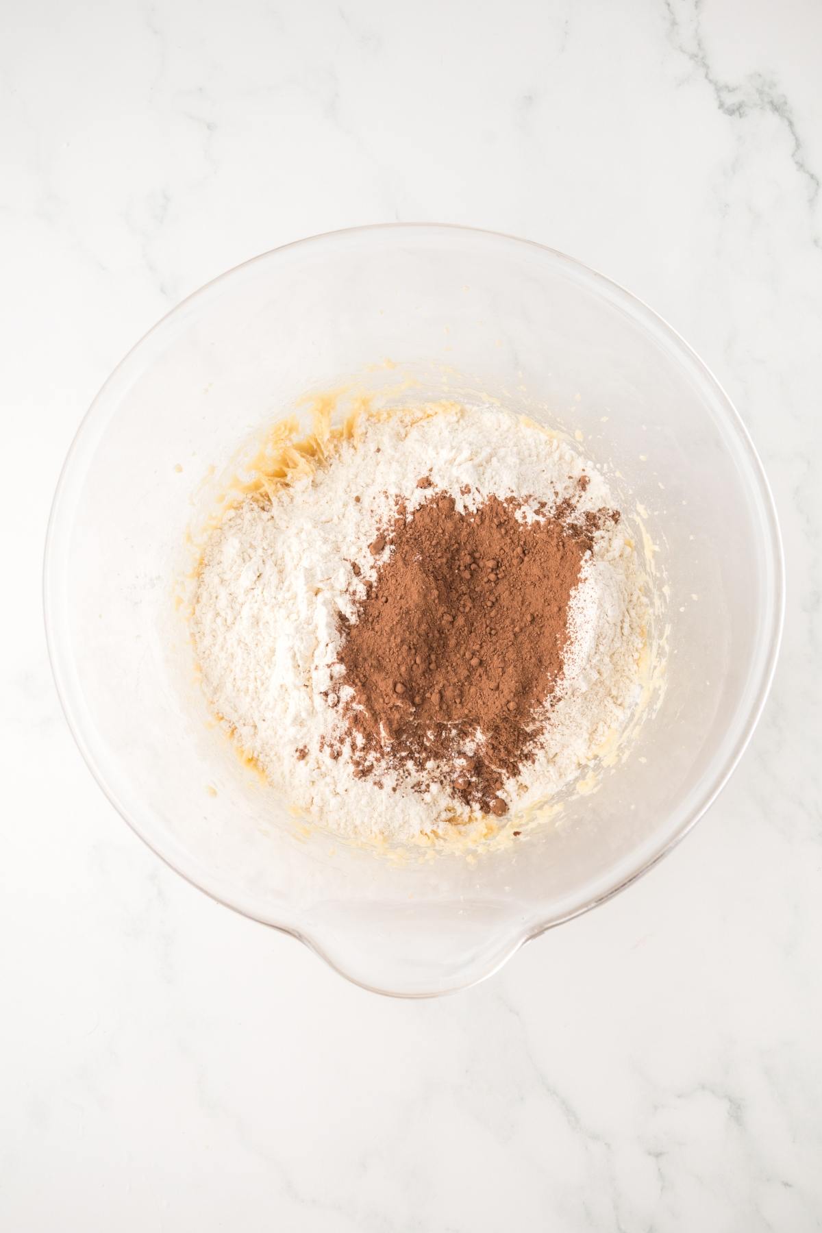 Clear mixing bowl with flour and cocoa powder on top of dough, on a white marble surface.