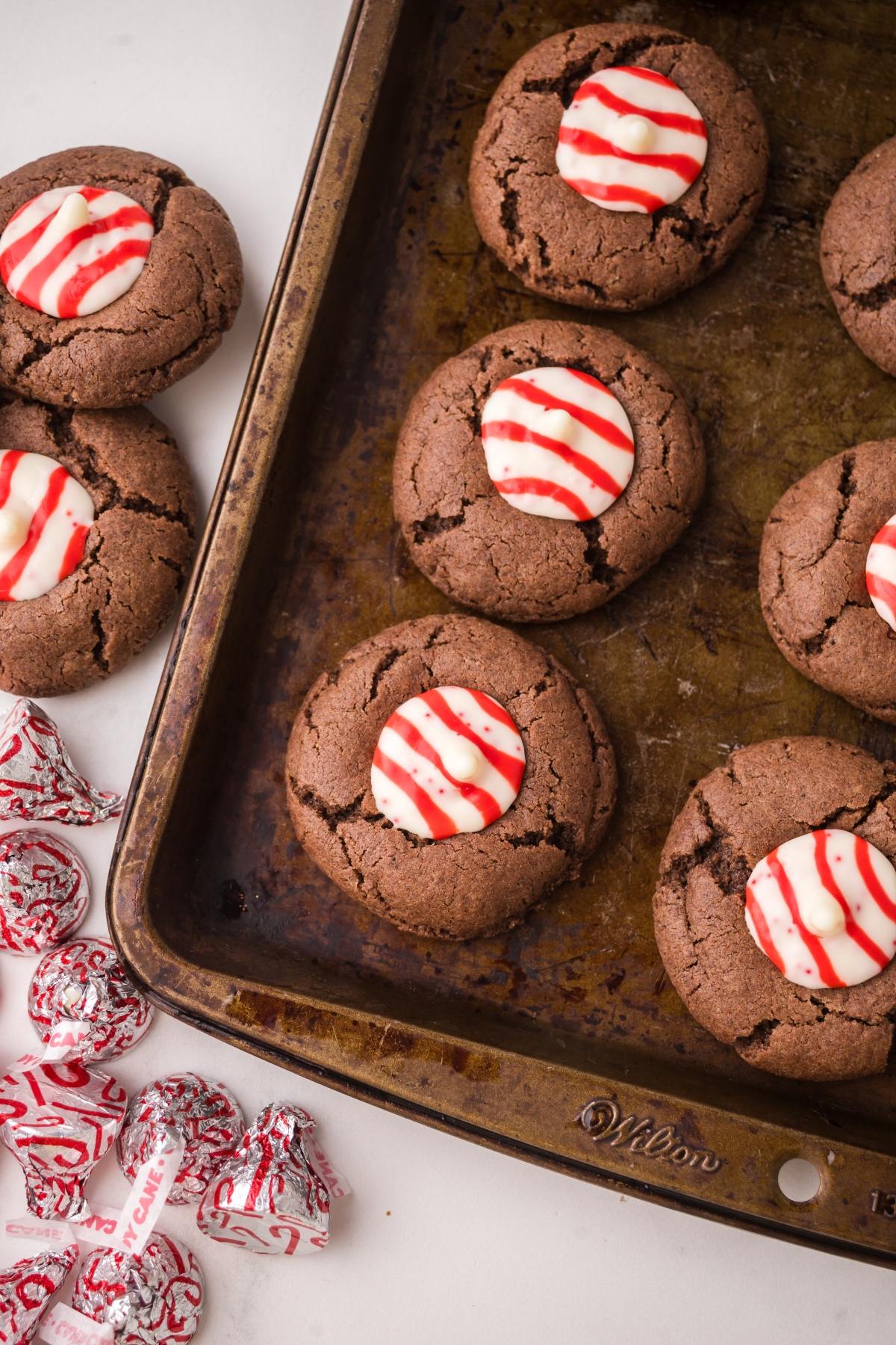 Chocolate Peppermint kiss cookies with red and white striped peppermint candies on a baking tray and nearby on a white surface.