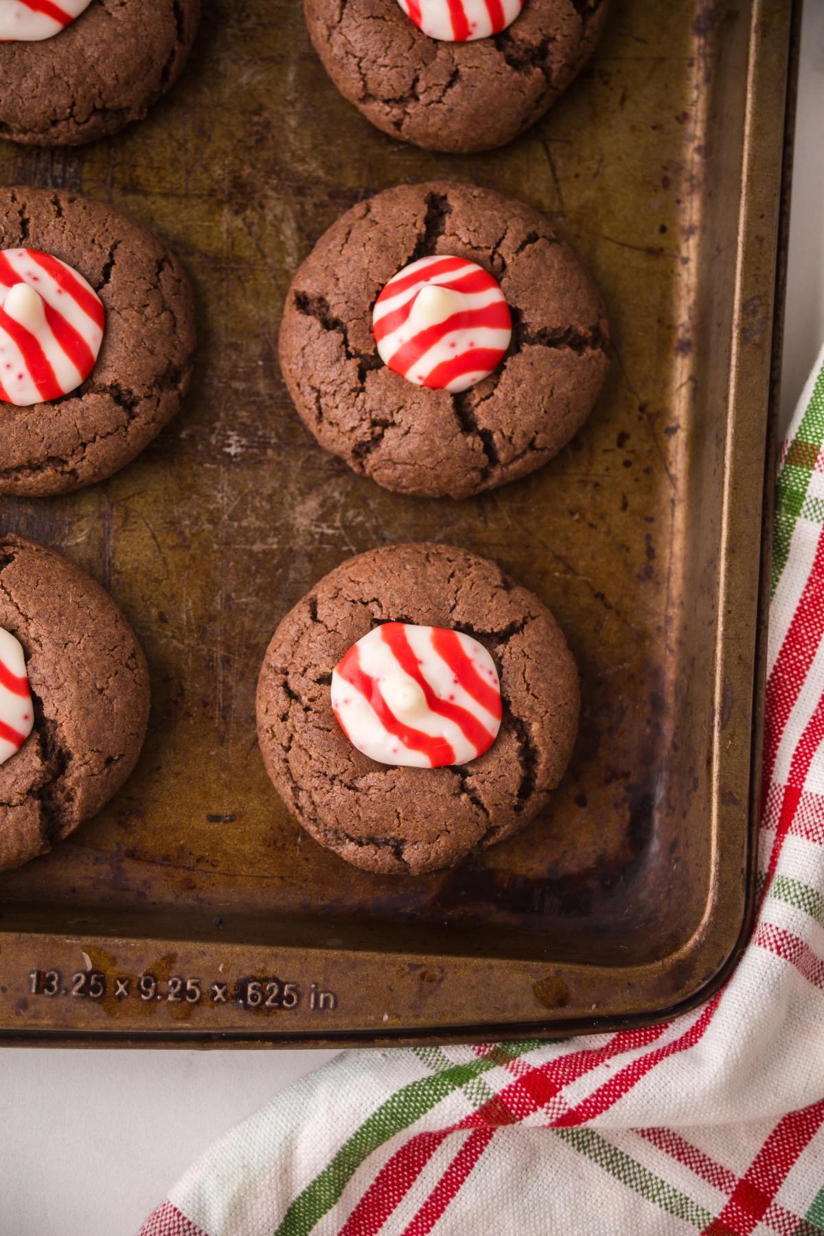 Chocolate Peppermint kiss cookies topped with red and white peppermint candies on a baking tray next to a striped cloth.