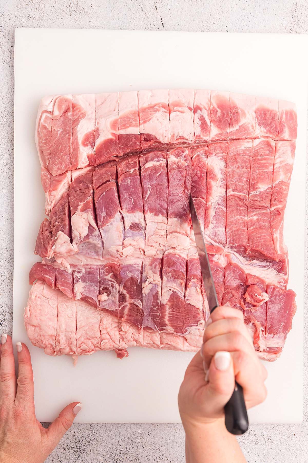 A hand holds a knife, scoring a large raw pork belly for porchetta roast on a white cutting board.