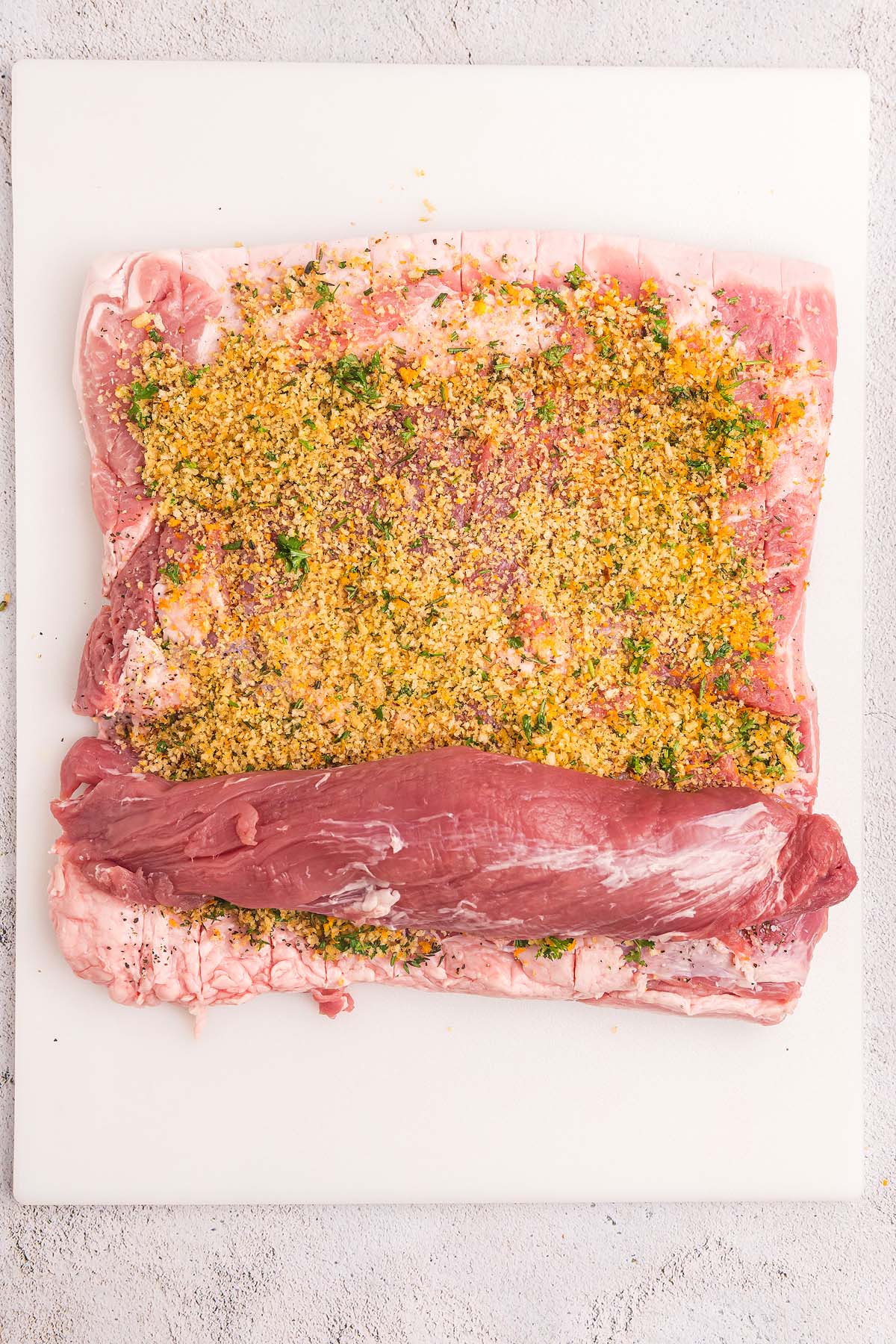 Raw pork loin prepped for a classic porchetta roast, topped with a savory breadcrumb and herb mixture, ready to be rolled and cooked on a white surface.
