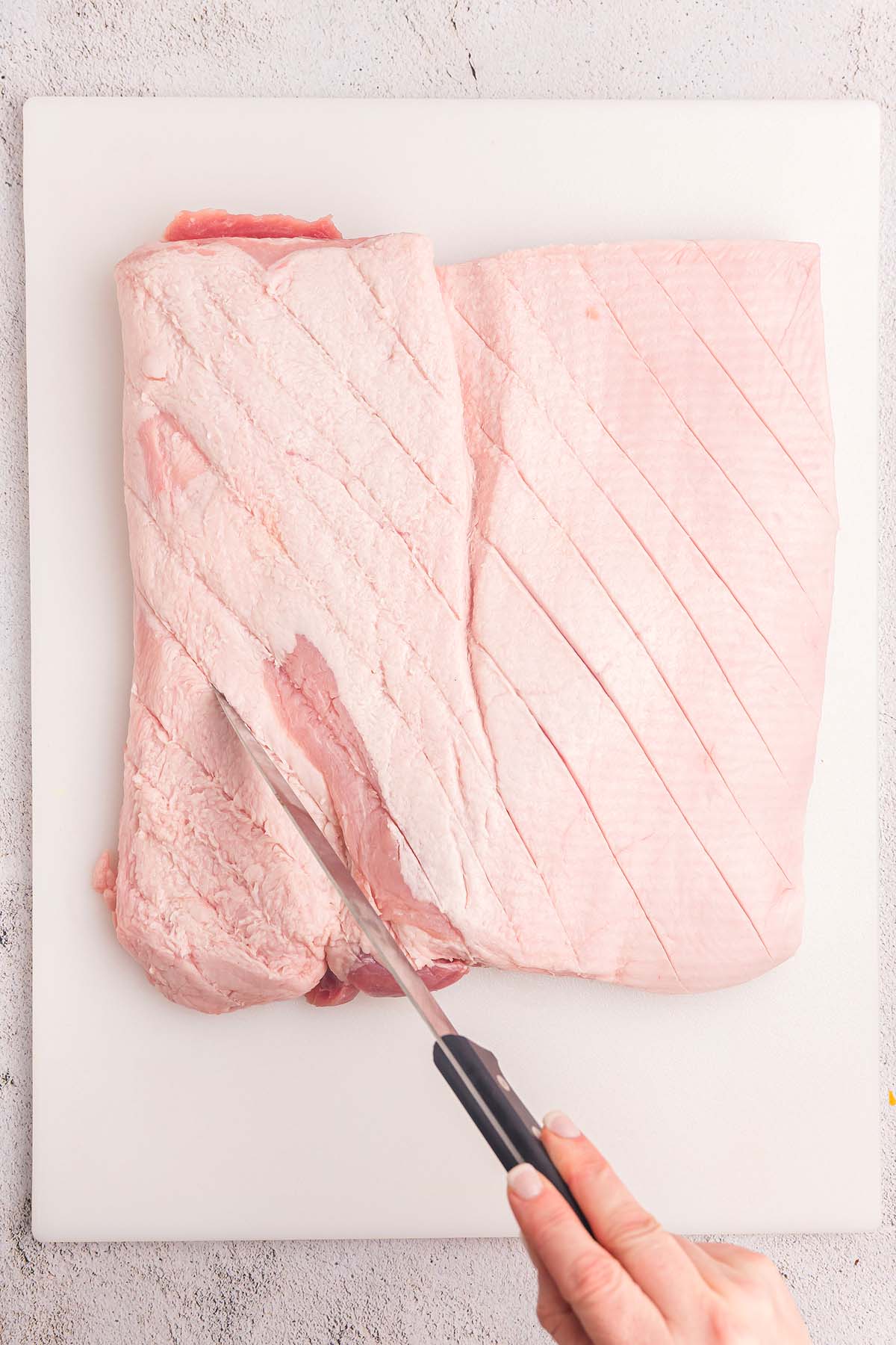 A hand scoring the fat on a large raw pork belly with a knife on a white cutting board, preparing it for a flavorful porchetta roast.