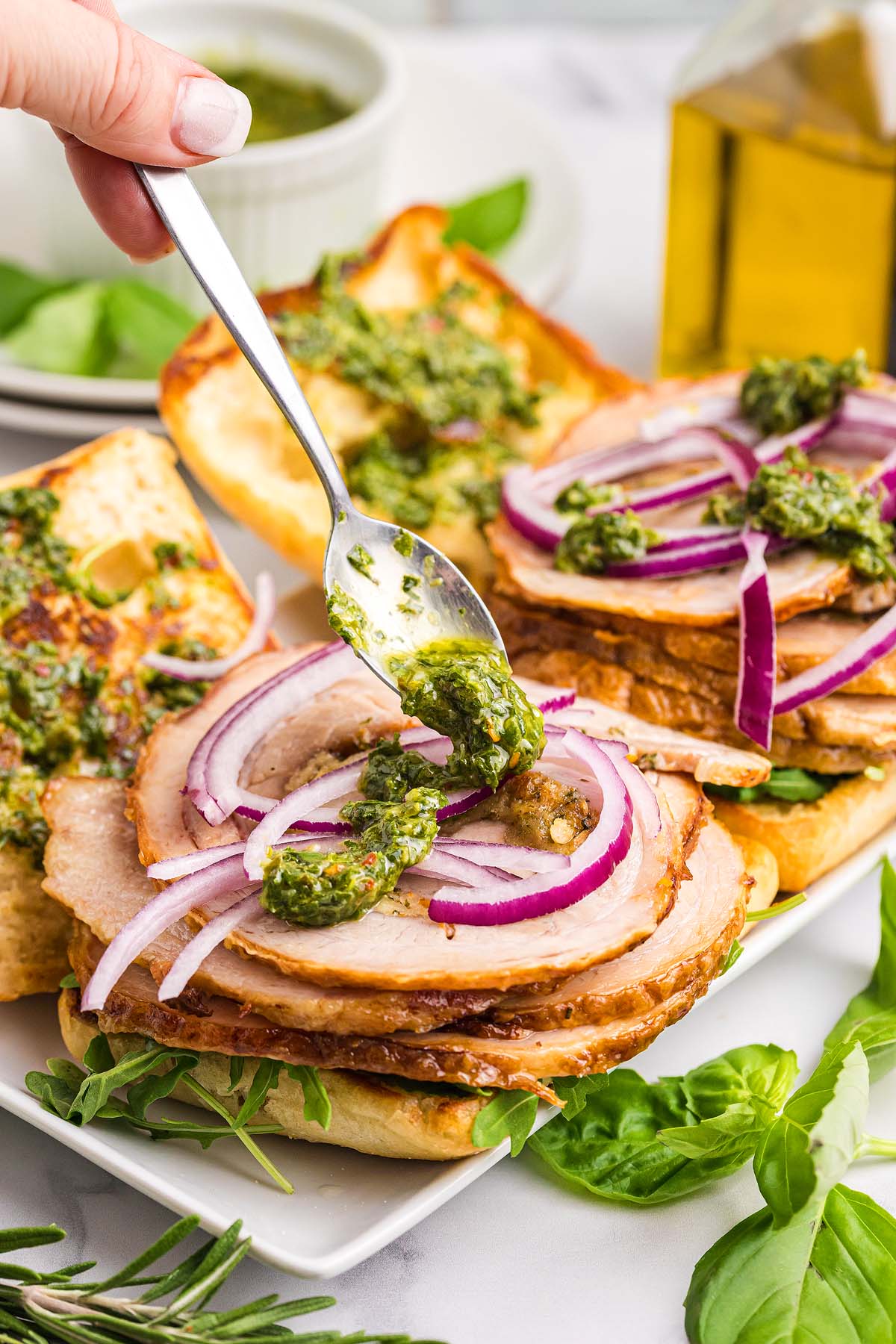 A hand spoons green pesto onto a Porchetta Sandwich layered with roast pork and red onion on toasted bread.