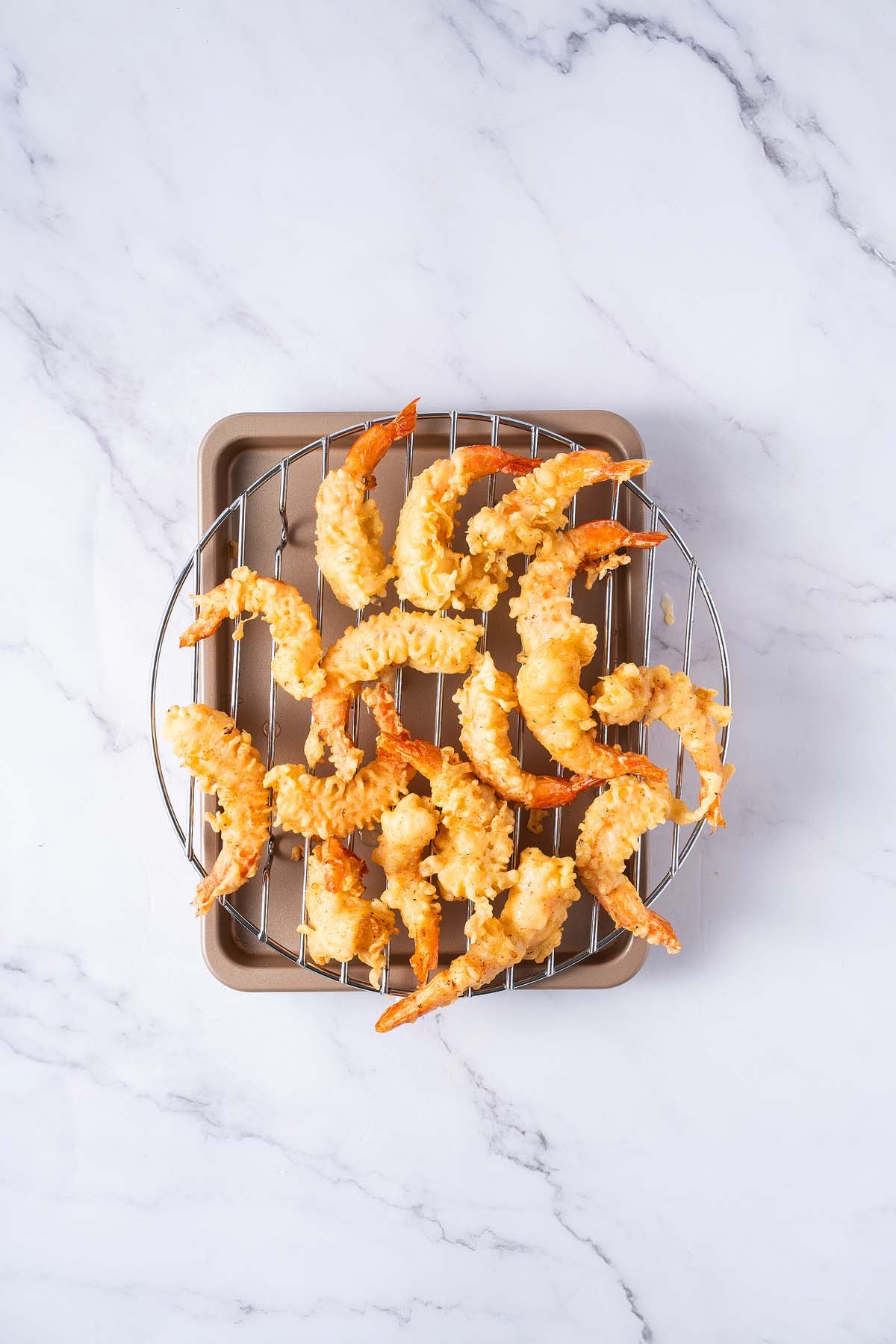Crispy shrimp tempura arranged on a wire rack over a baking tray, set against a white marble surface.
