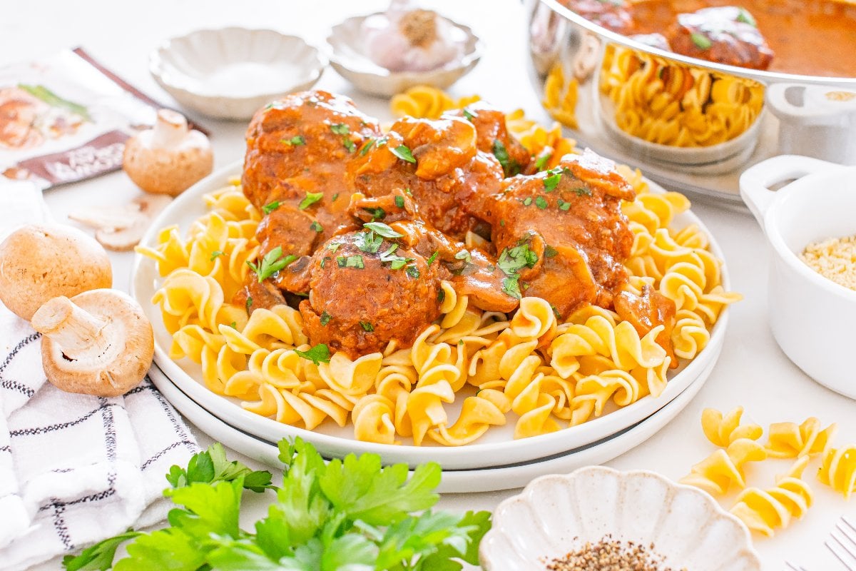 A plate of egg noodles topped with Salisbury Steak Meatballs in tomato sauce, surrounded by fresh herbs and mushrooms.