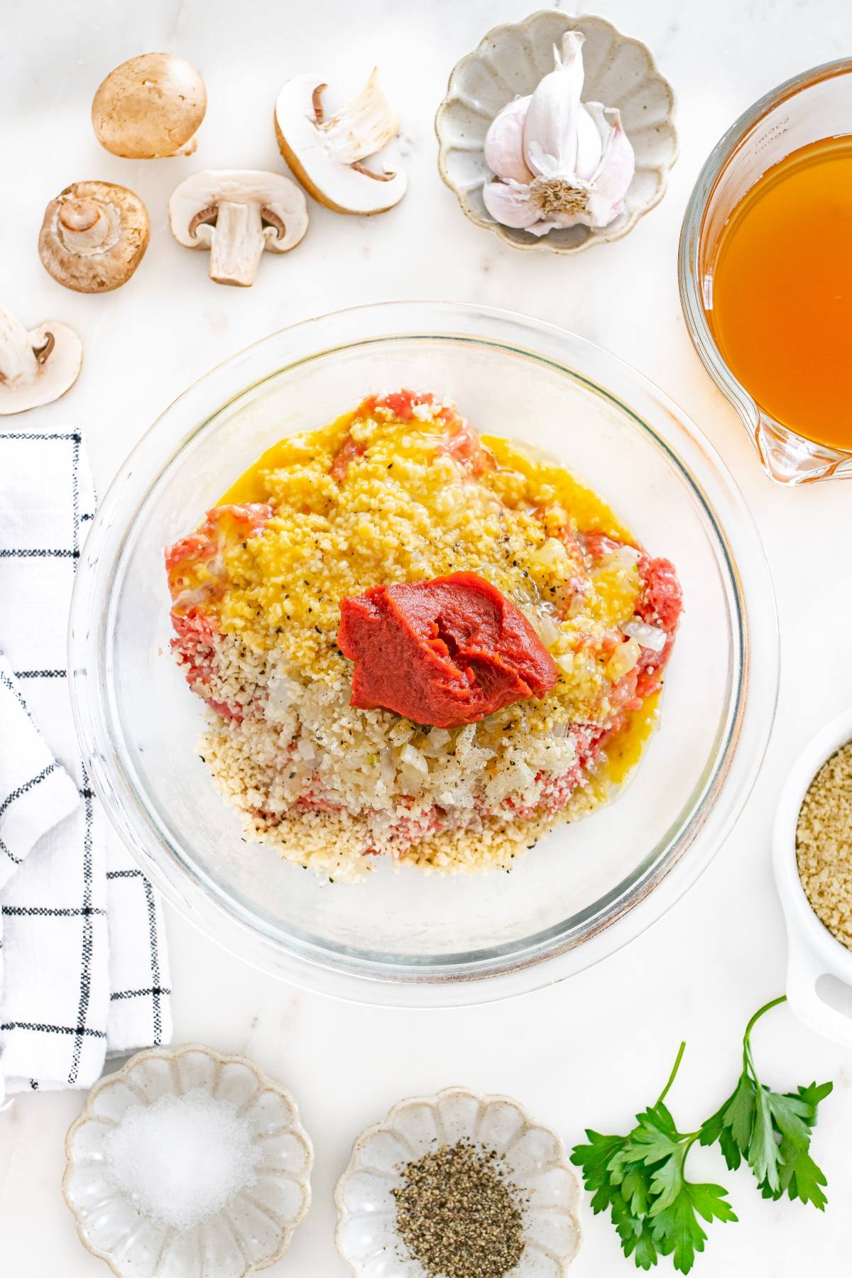 Glass bowl with ground meat, breadcrumbs, egg, tomato paste, and seasonings, surrounded by cooking ingredients.