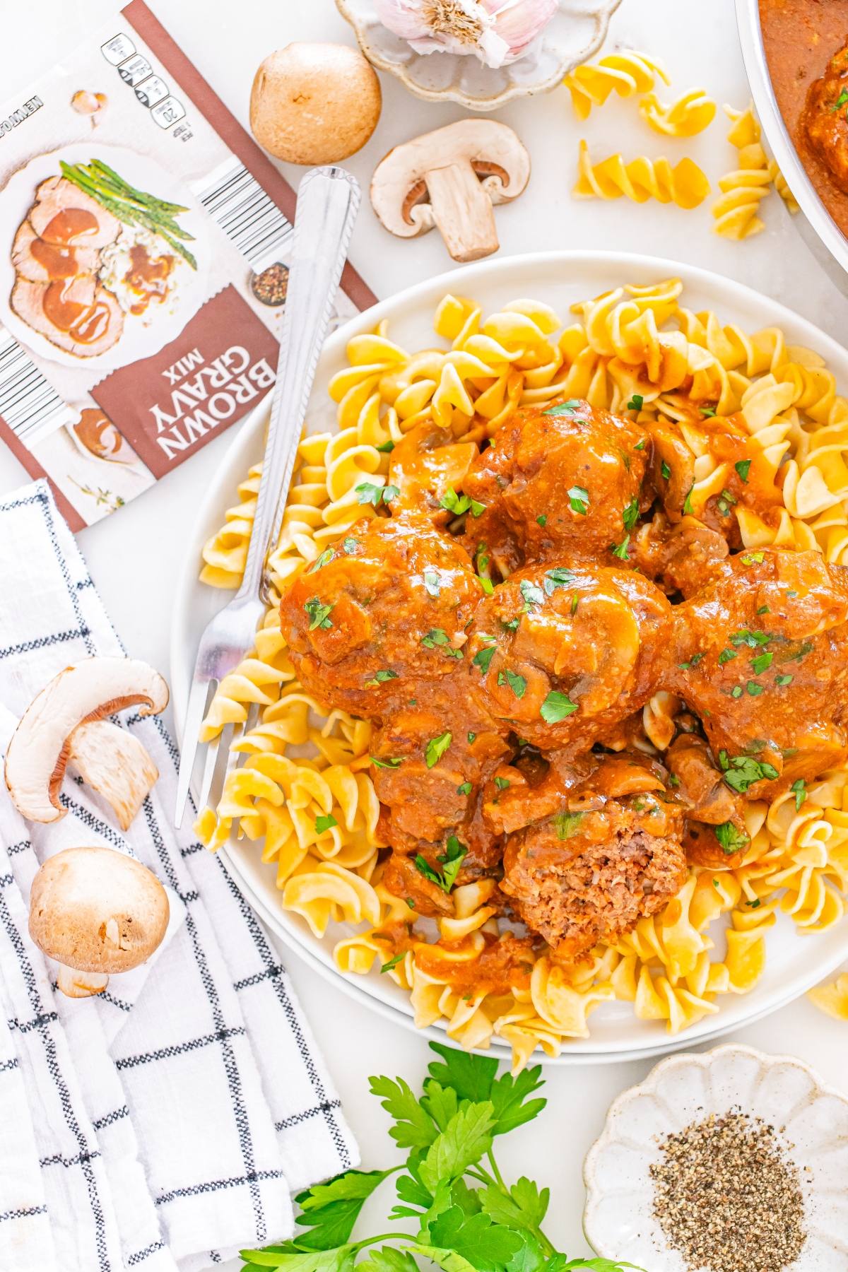 Plate of meatballs in brown gravy over egg noodles, surrounded by mushrooms, herbs, and packaged gravy mix.