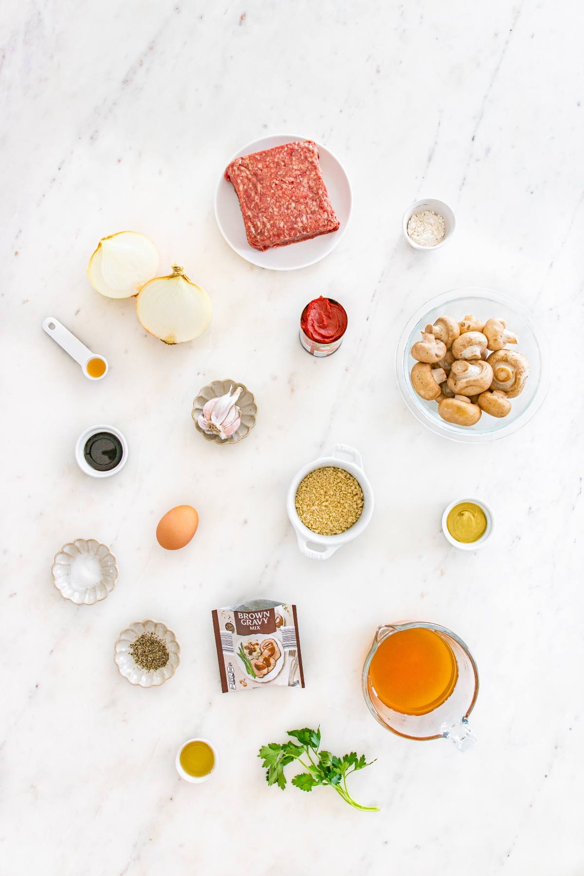 Ingredients for a recipe laid out on a white surface, including ground meat, mushrooms, onion, egg, and seasonings.