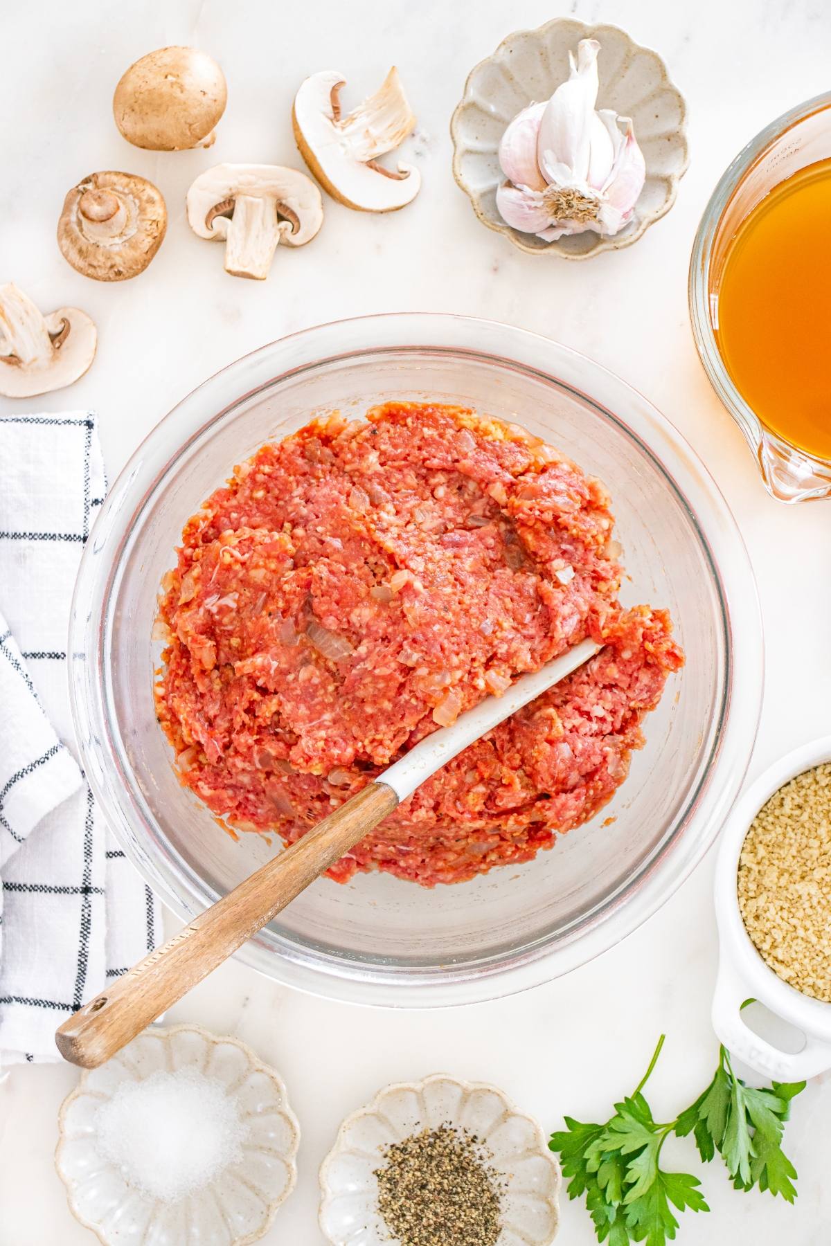 Bowl of raw ground meat mixture with a wooden spoon, surrounded by mushrooms, garlic, broth, and seasonings.