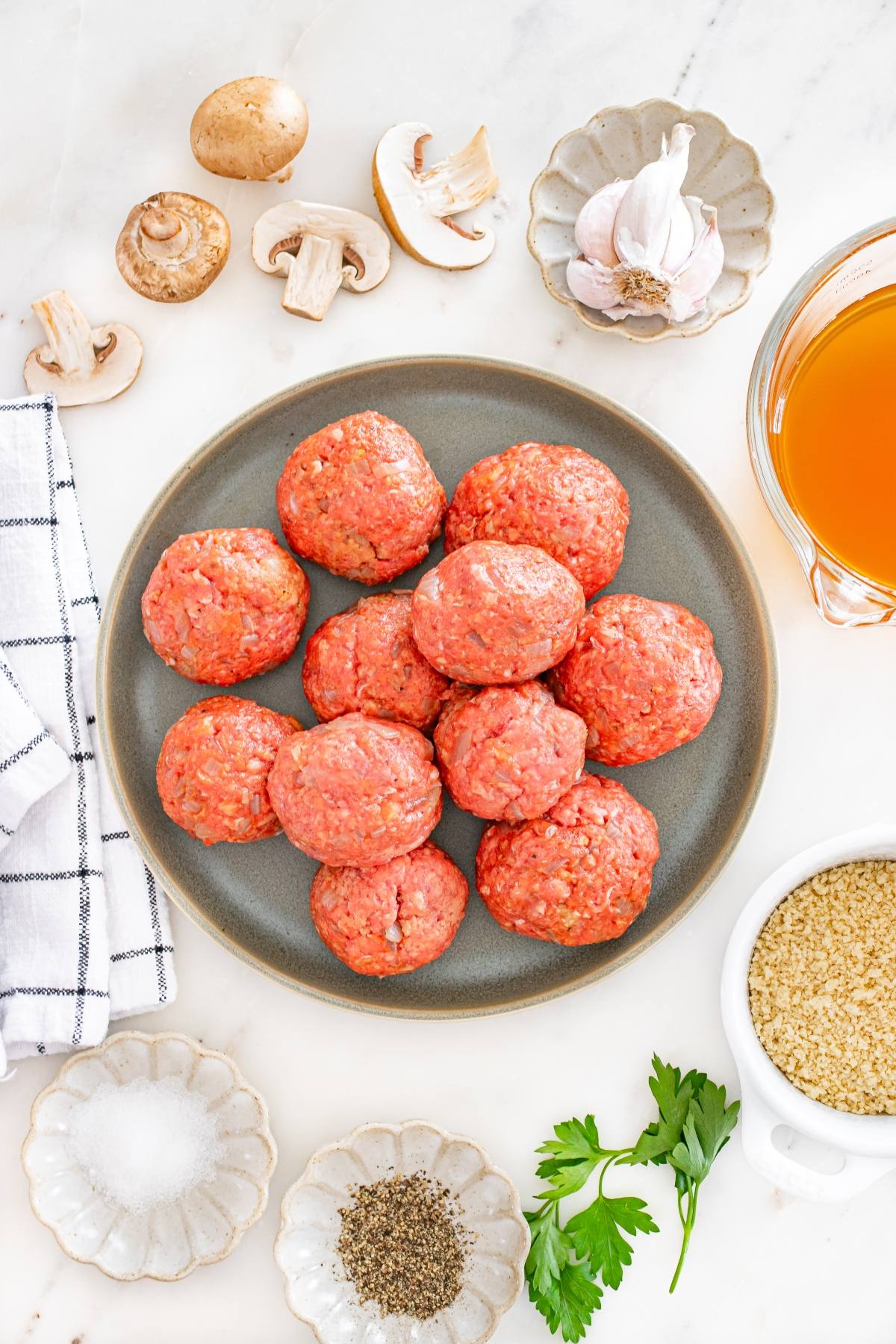 Raw meatballs on a plate, surrounded by mushrooms, garlic, broth, parsley, seasonings, and quinoa on a marble surface.