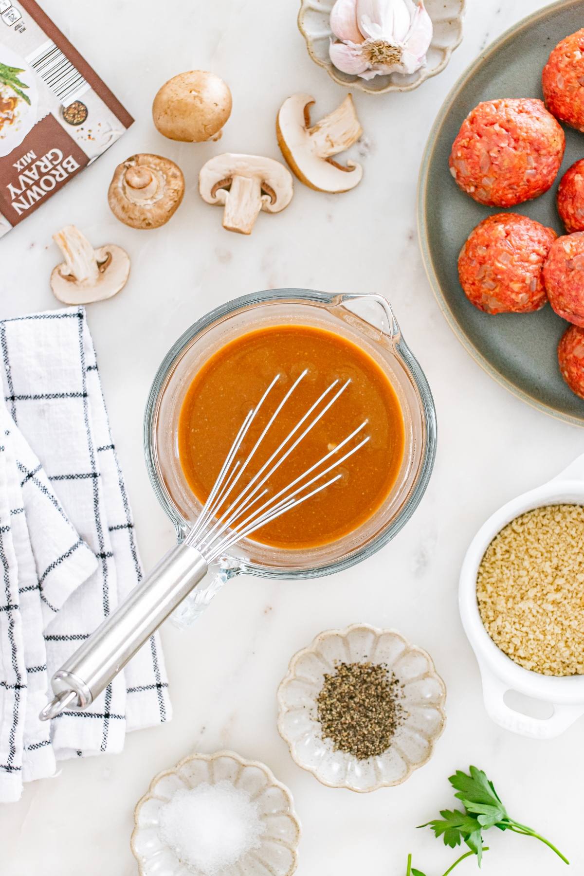 Whisk in bowl of brown gravy, raw meatballs, mushrooms, seasonings, breadcrumbs, and herbs on a marble counter.