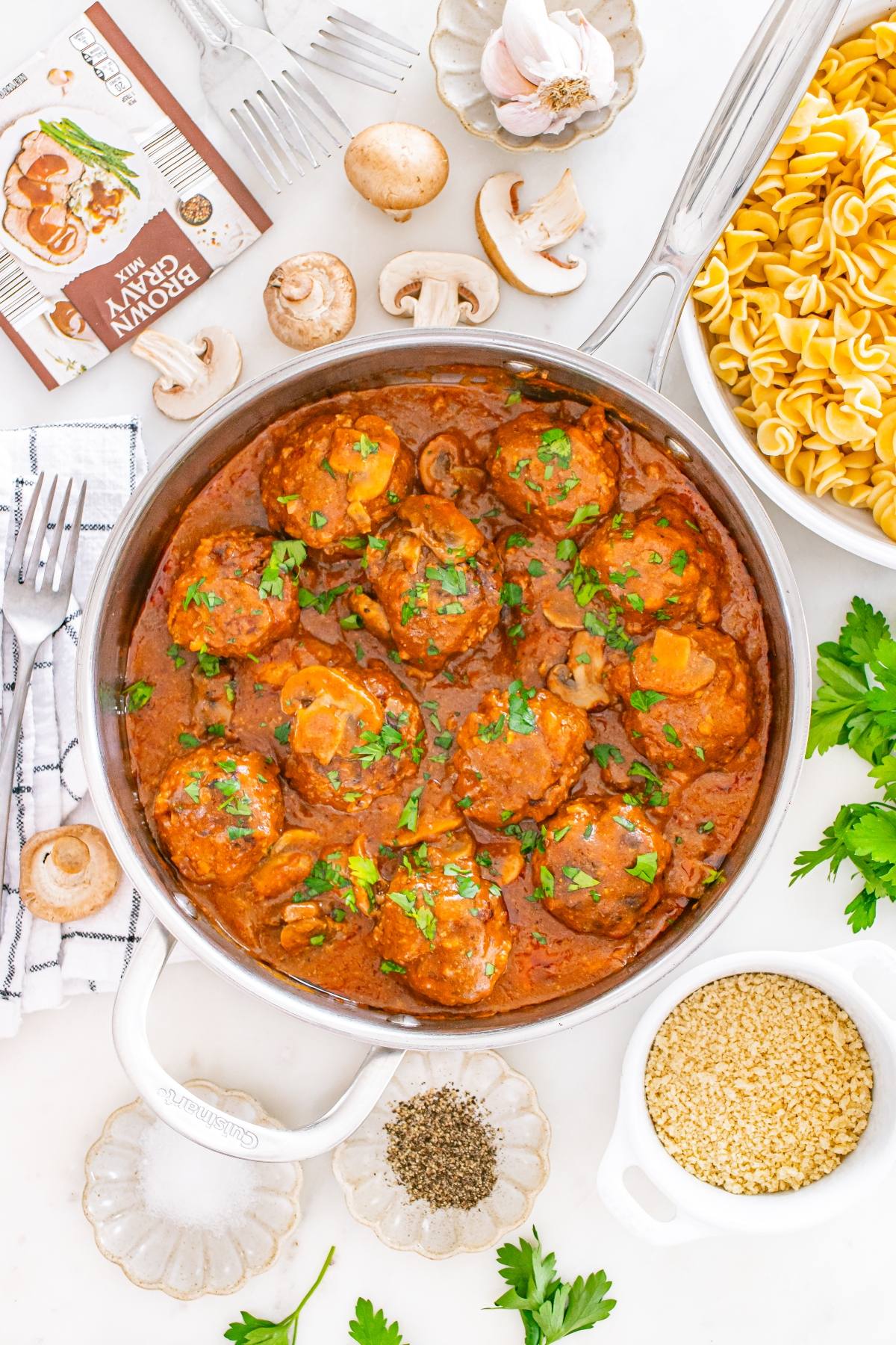 A skillet of meatballs in tomato sauce with mushrooms, surrounded by pasta, spices, herbs, and garlic.