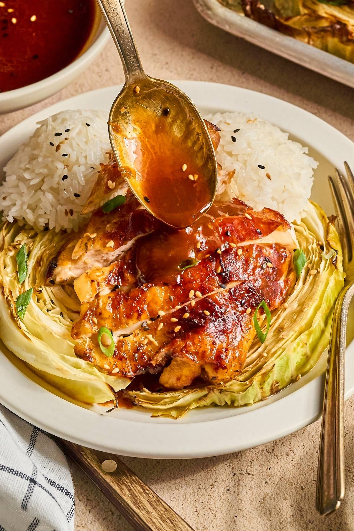 A spoon drizzles sauce over glazed chicken, rice, and cabbage on a white plate, garnished with sesame seeds.