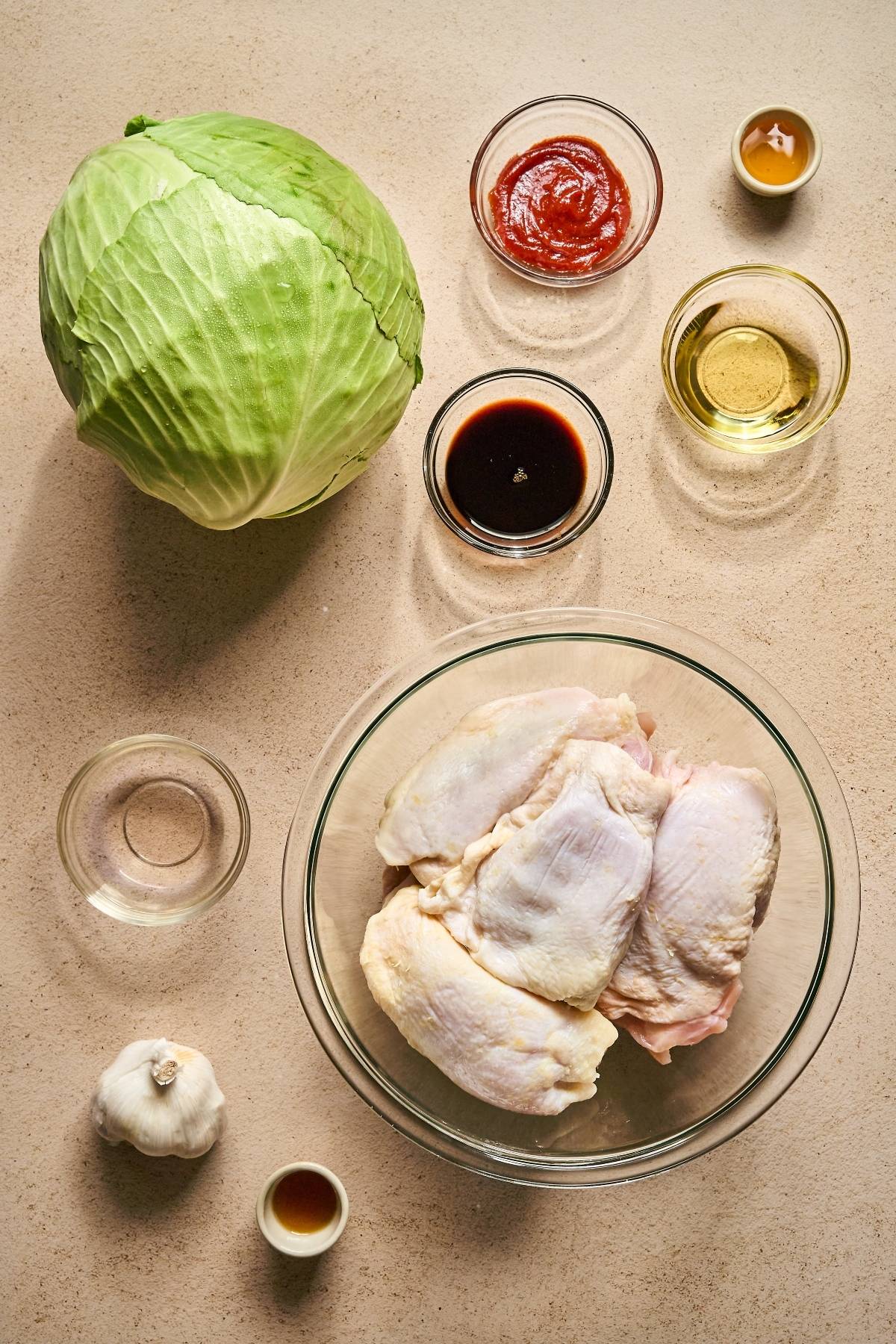 Raw chicken, a head of cabbage, garlic, and sauces in bowls arranged on a beige surface for cooking prep.