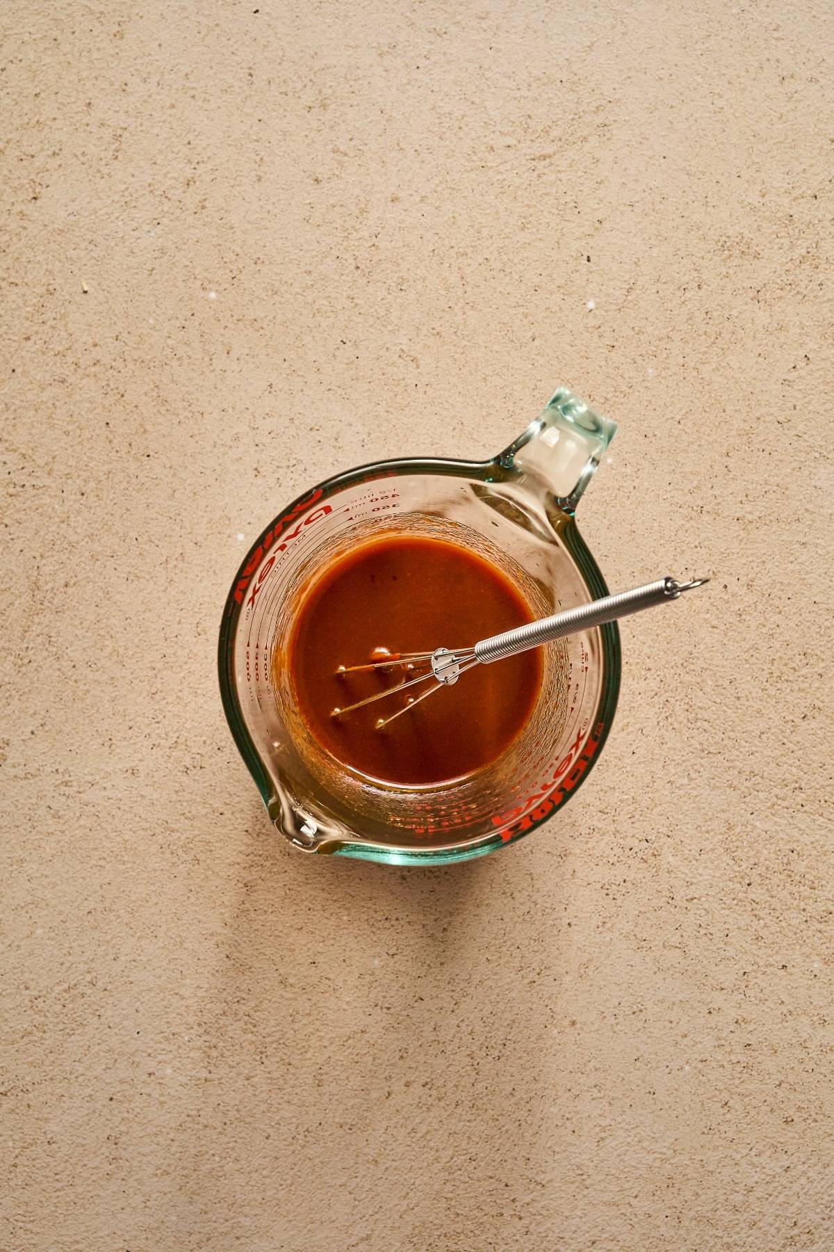 Glass measuring cup with brown sauce and a small whisk on a light tan surface.