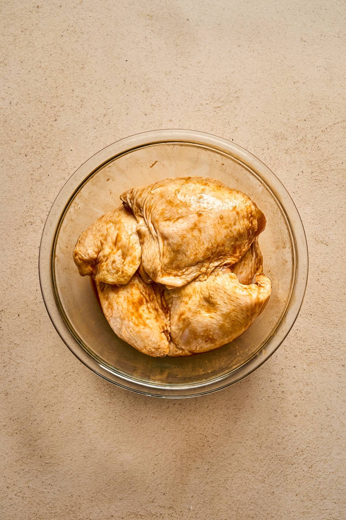 Raw, marinated chicken breasts in a clear glass bowl on a beige surface, viewed from above.