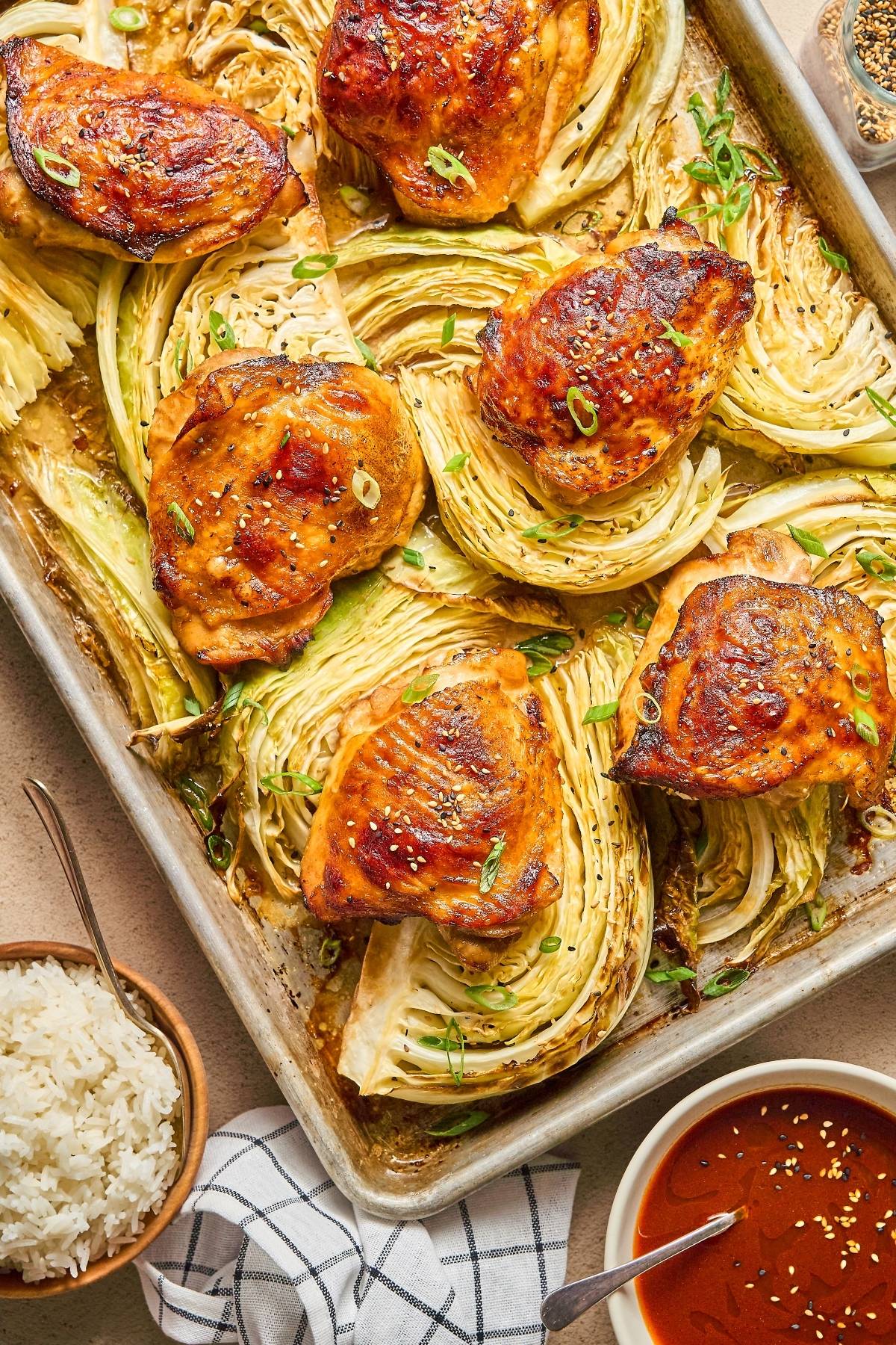 Roasted chicken thighs on a baking sheet with cabbage wedges, served with rice and sauce on the side.