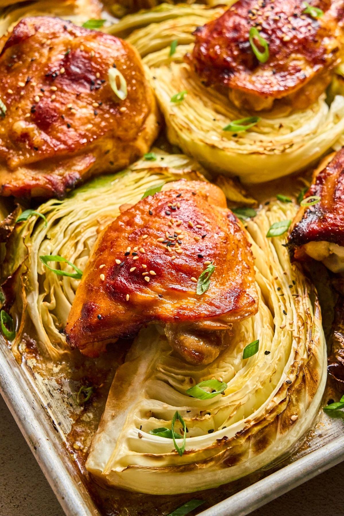 Roasted chicken thighs and cabbage wedges garnished with sesame seeds and sliced green onions on a baking sheet.