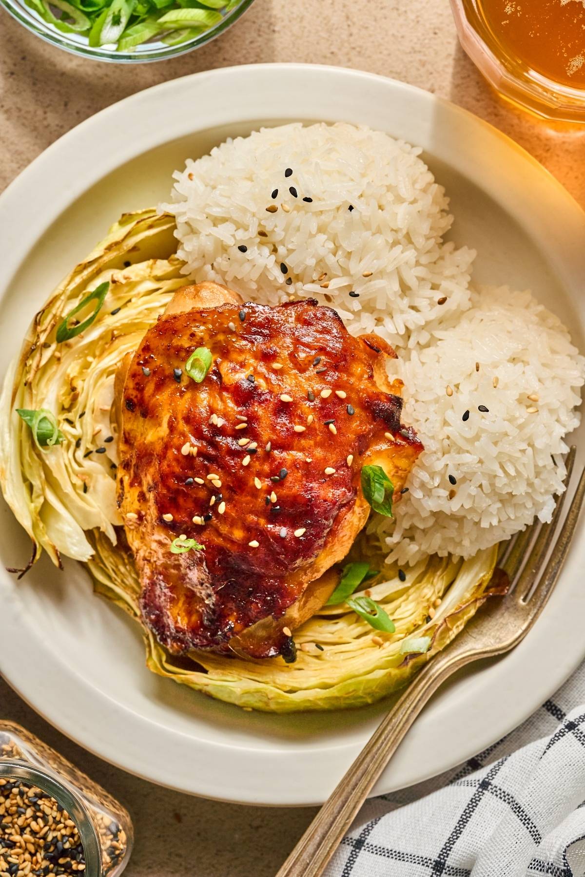 A plate of glazed chicken thigh, roasted cabbage, and two scoops of white rice sprinkled with sesame seeds.