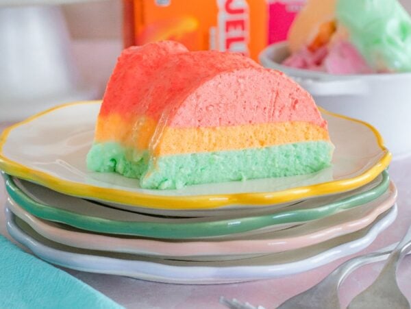 A slice of colorful Sherbet Layered Jello Mold sits on a stack of plates with forks beside it.