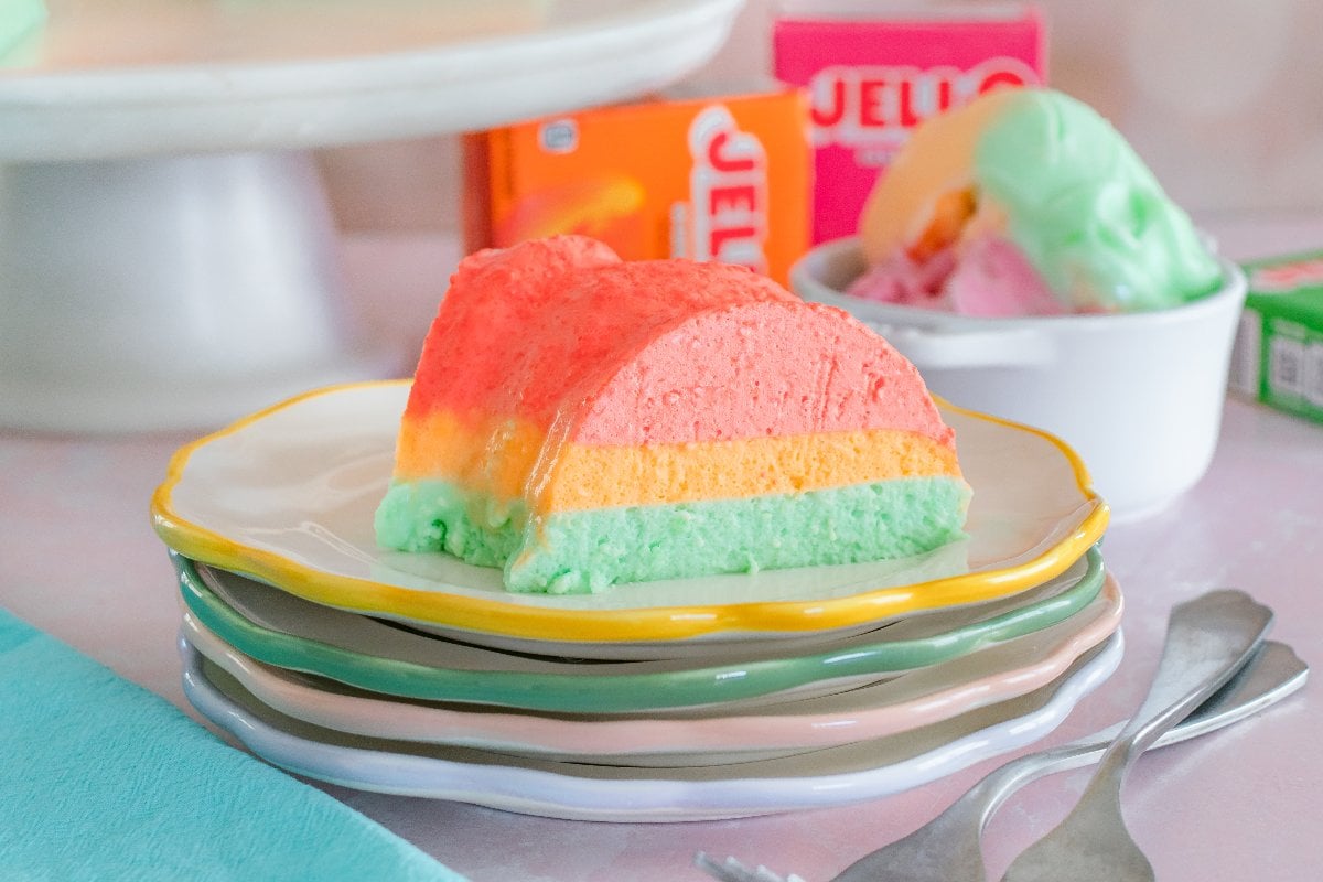 A colorful Sherbet Layered Jello Mold dessert slice on stacked plates, with Jello mix boxes and ice cream in the background.