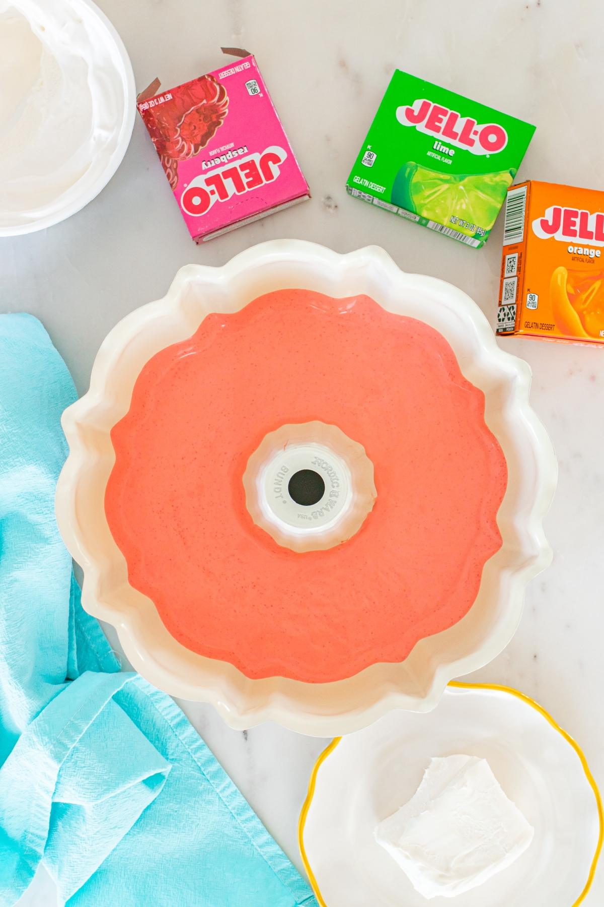 Bundt pan with pink Jello mixture, Jello boxes, whipped topping, and cream cheese on a counter.