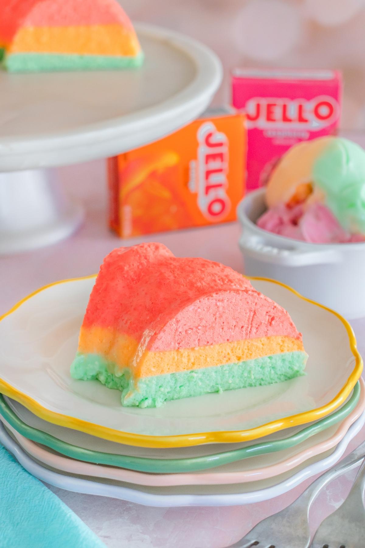 A slice of layered pastel Jello dessert on a plate, with Jello boxes and ice cream in the background.