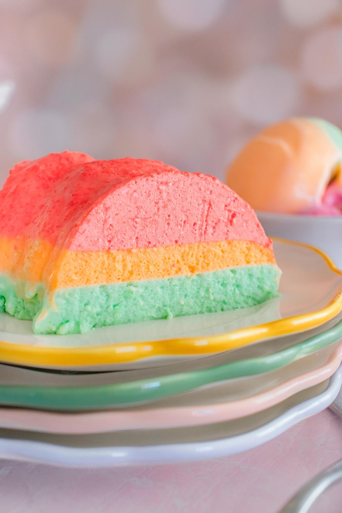 A slice of layered pastel mousse dessert in pink, orange, and green on a stack of colorful plates.