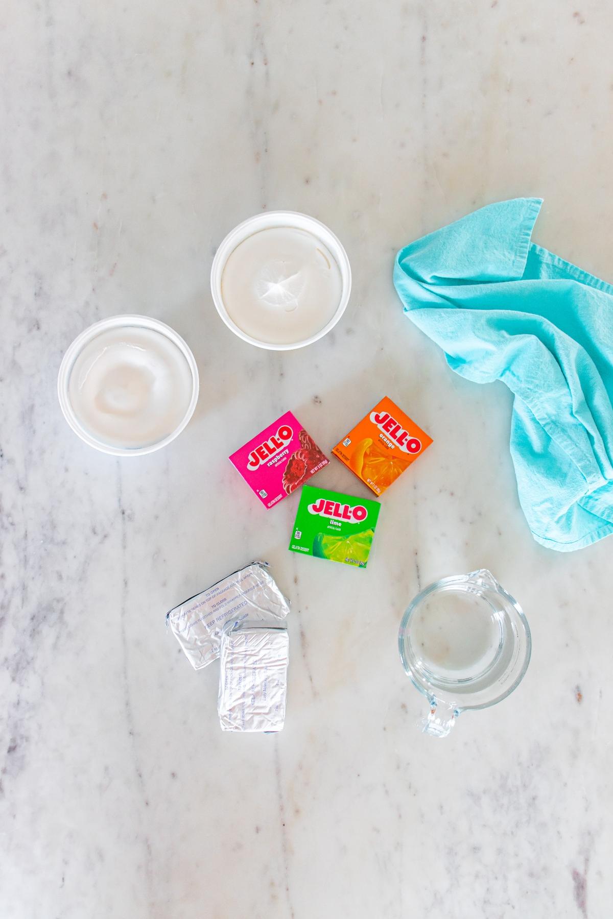 Two bowls of whipped topping, three Jell-O boxes, cream cheese, a measuring cup, and a blue cloth on a counter.