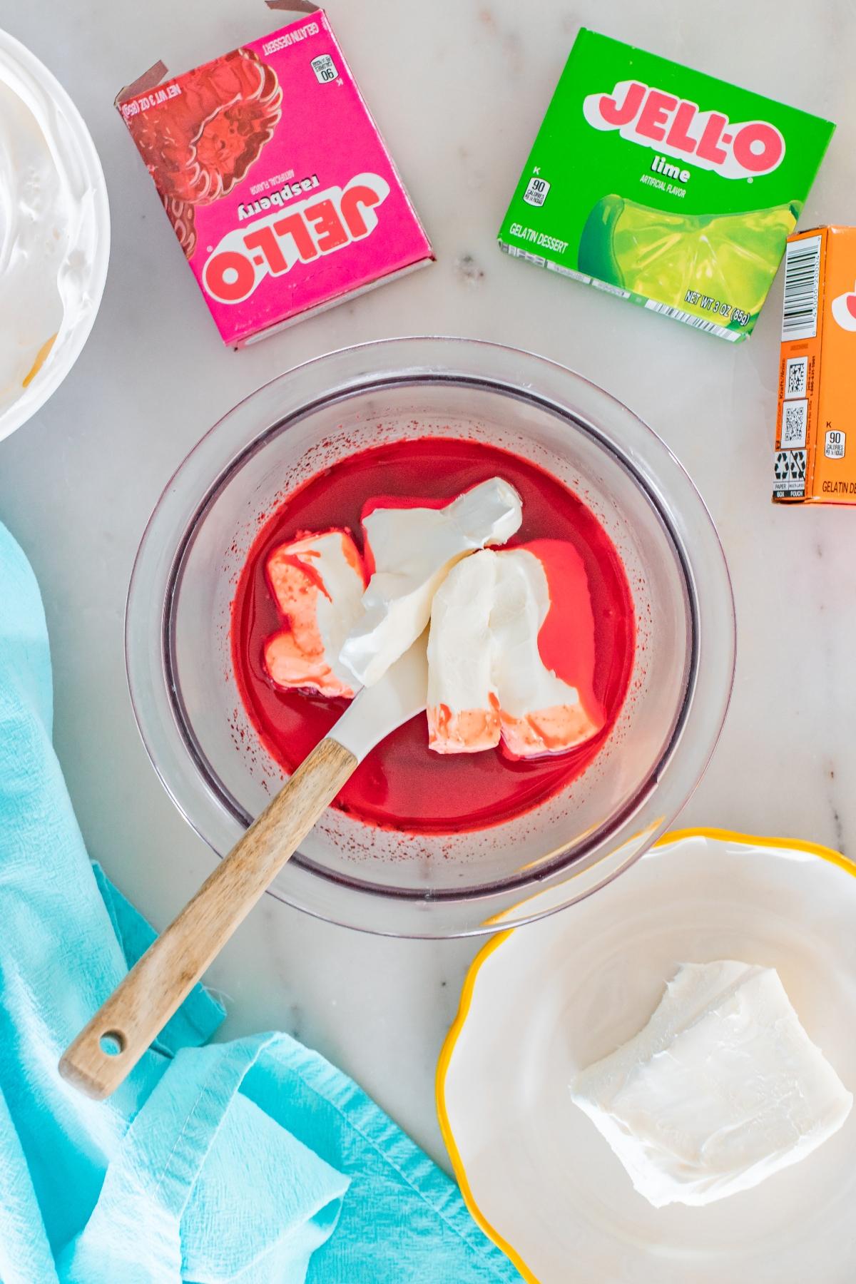 A bowl of red Jello mix with whipped topping being stirred, surrounded by Jello boxes and cream cheese.