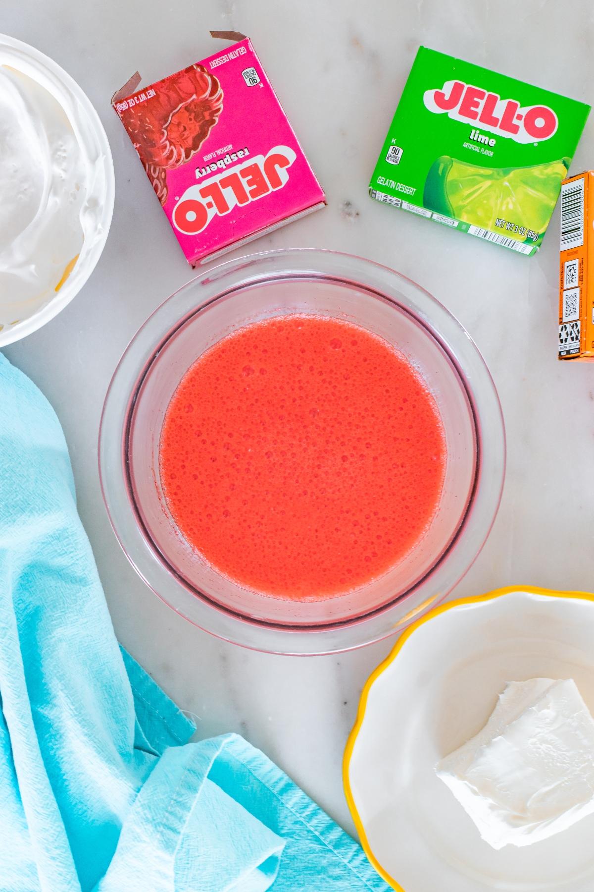 A bowl of red liquid, Jello boxes, whipped topping, cream cheese, and a blue cloth on a white surface.