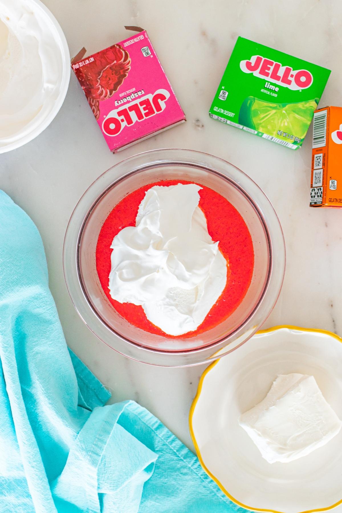 A bowl with red gelatin and whipped topping, surrounded by Jell-O boxes and a blue towel on a white surface.