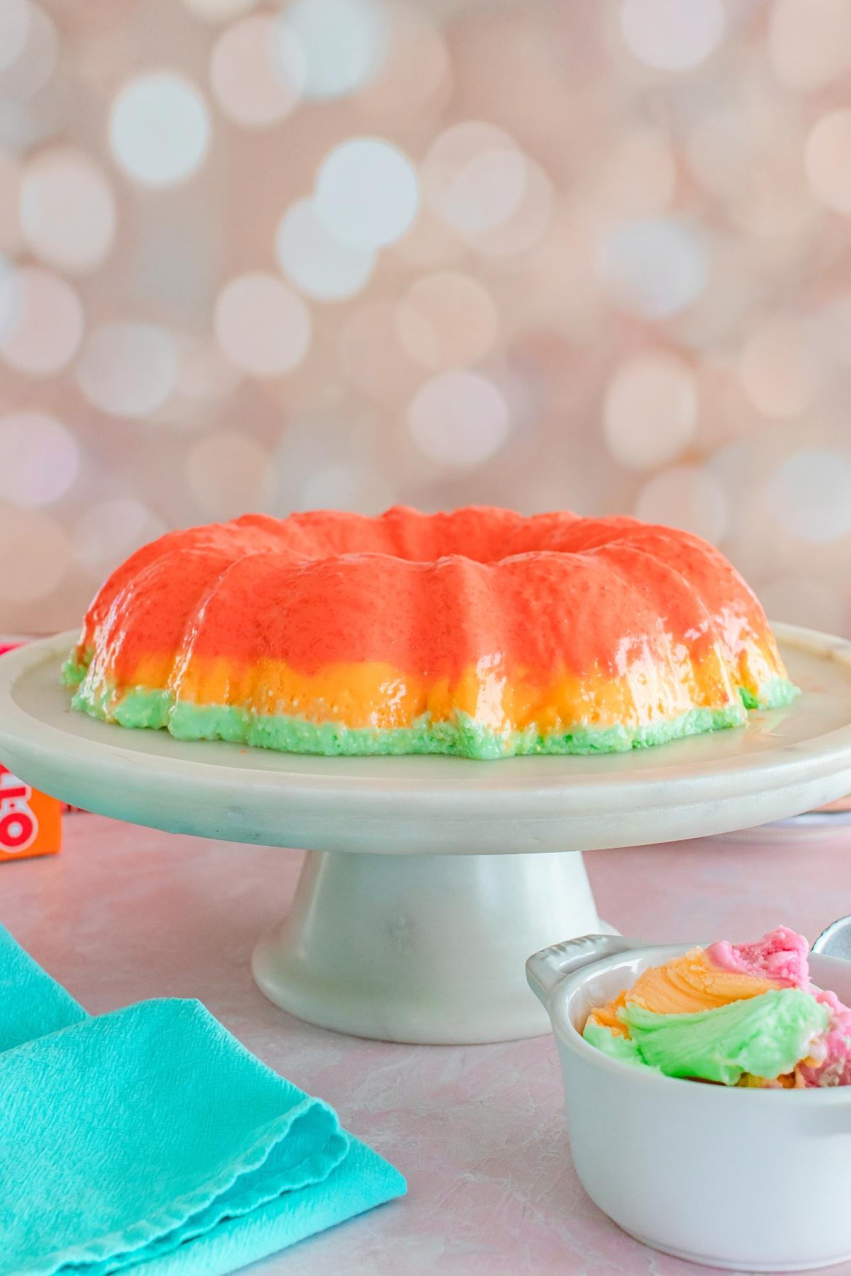 A colorful layered gelatin mold on a cake stand, with a teal napkin and pastel swirl ice cream nearby.