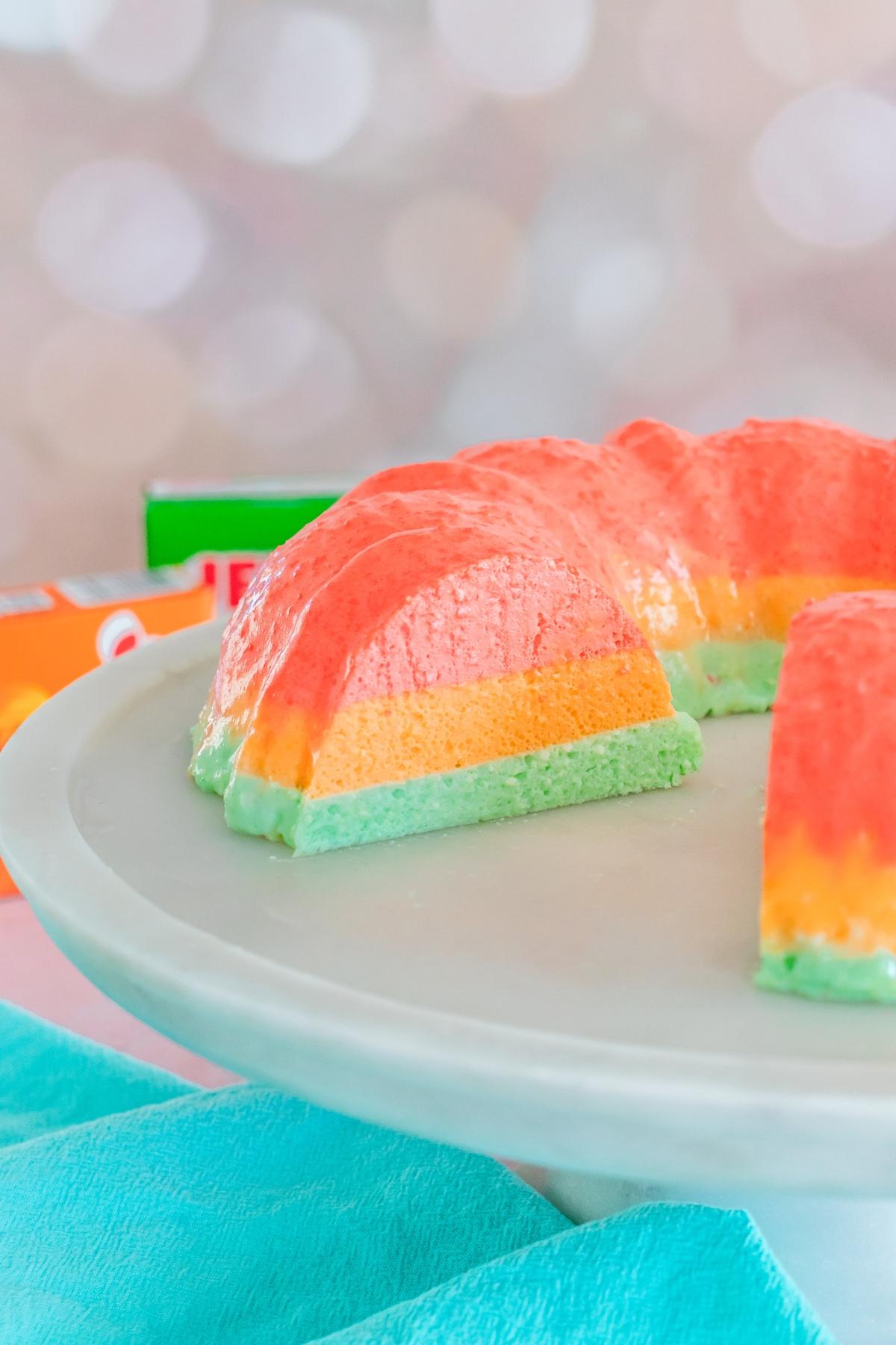 A colorful, layered gelatin dessert with pink, orange, and green sections on a white cake stand.