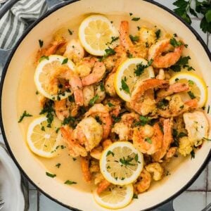 Cooked Shrimp Francese with lemon slices and parsley in a pan, surrounded by fresh bread, lemon, and herbs on a table.