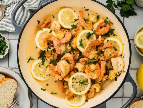 Cooked Shrimp Francese with lemon slices and parsley in a pan, surrounded by fresh bread, lemon, and herbs on a table.