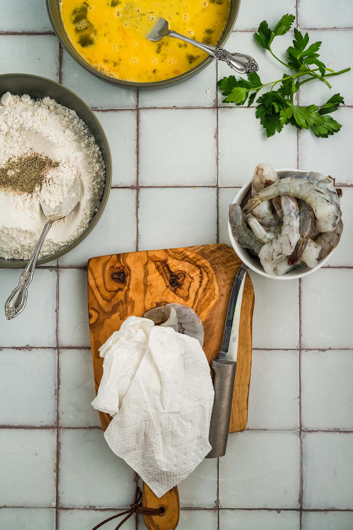 Shrimp, flour, eggs, parsley, and a knife on a cutting board, prepped for cooking Shrimp Francese on a tiled surface.