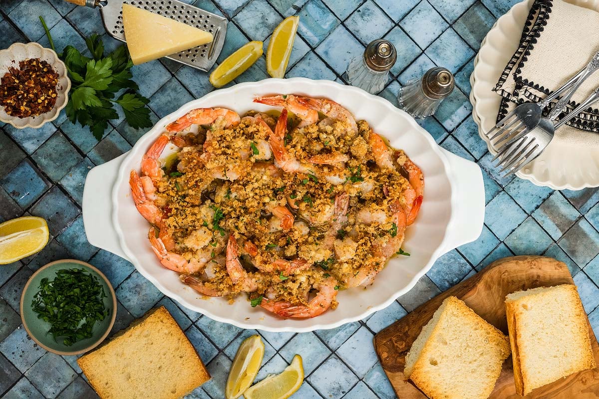 Shrimp Oreganata baked with crispy breadcrumbs in a white dish, surrounded by bread slices, lemon wedges, and fresh herbs.