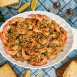 Shrimp Oreganata baked with golden breadcrumbs in a white dish, surrounded by lemon wedges, parsley, and slices of bread.