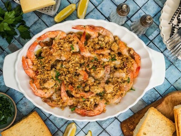 Shrimp Oreganata baked with golden breadcrumbs in a white dish, surrounded by lemon wedges, parsley, and slices of bread.