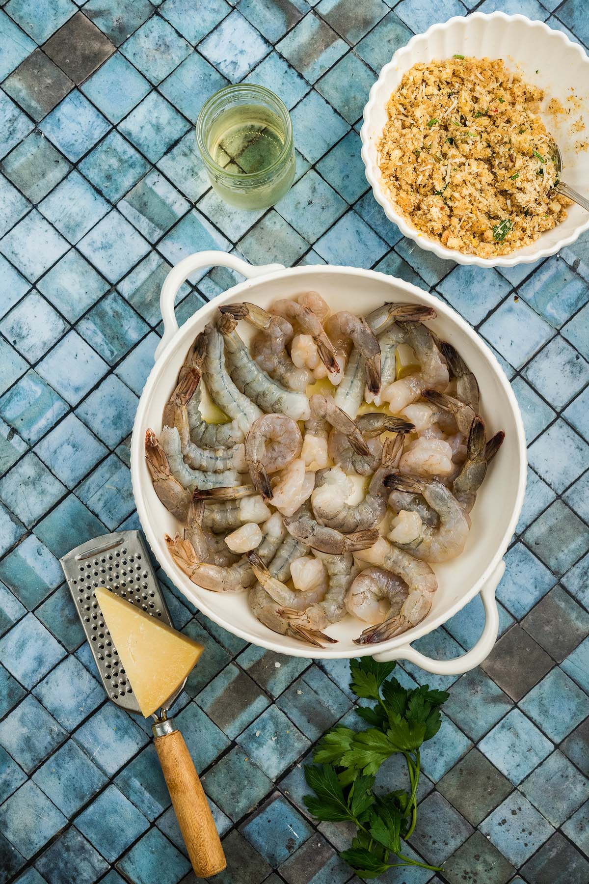 Raw shrimp in a white bowl, grated cheese, a jar, breadcrumbs in a dish, and parsley on a blue tiled surface—perfect ingredients for preparing Shrimp Oreganata.