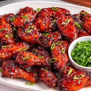 A platter of slow cooker spicy Korean wings glazed to perfection, garnished with sesame seeds and green onions, served with a bowl of sliced green onions.