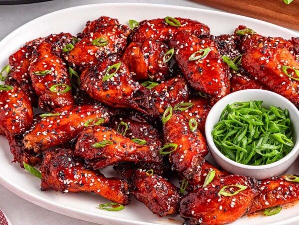 A platter of slow cooker spicy Korean wings glazed to perfection, garnished with sesame seeds and green onions, served with a bowl of sliced green onions.