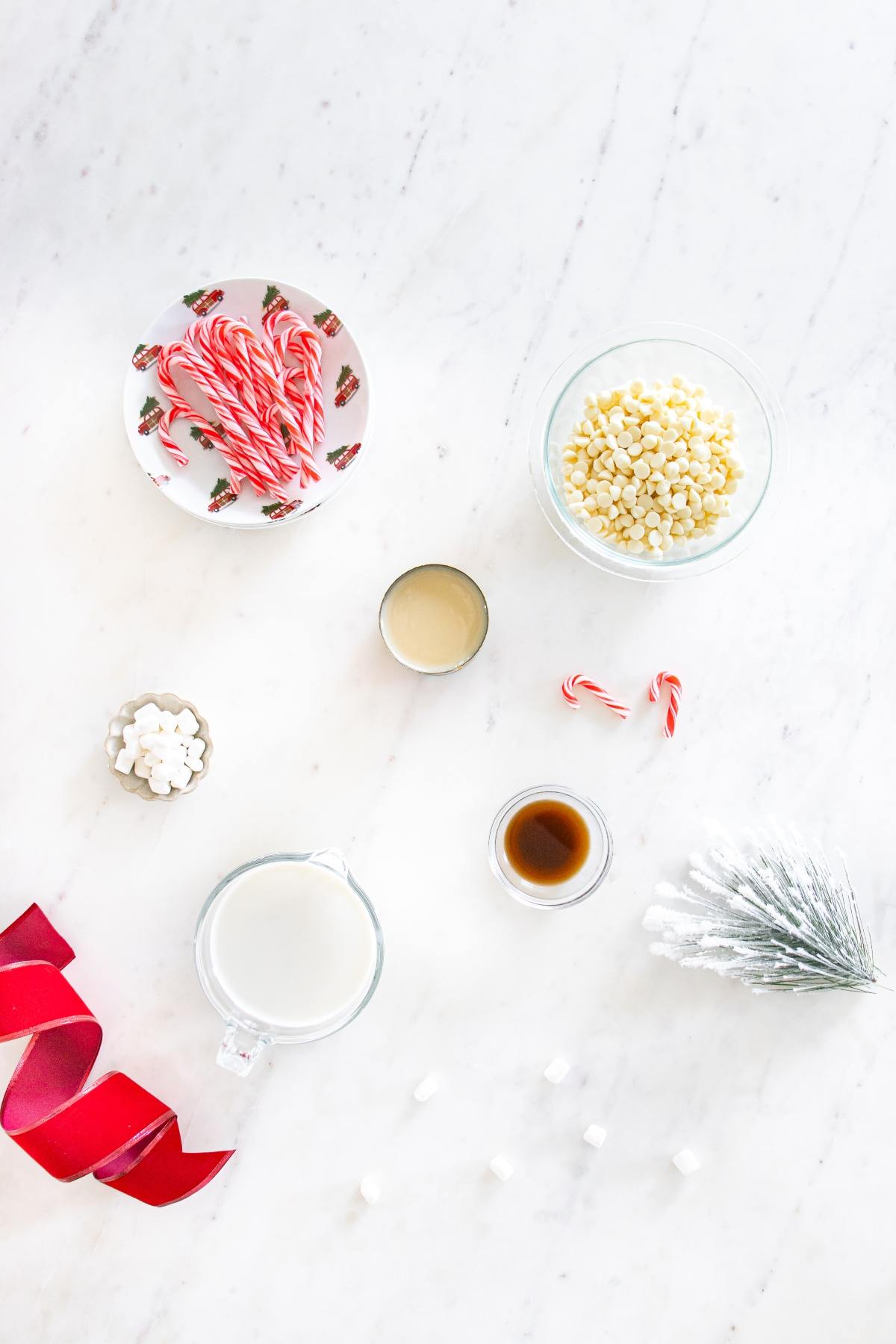 Candy canes, white chocolate chips, milk, marshmallows, and vanilla on a white surface with ribbon and pine decor.